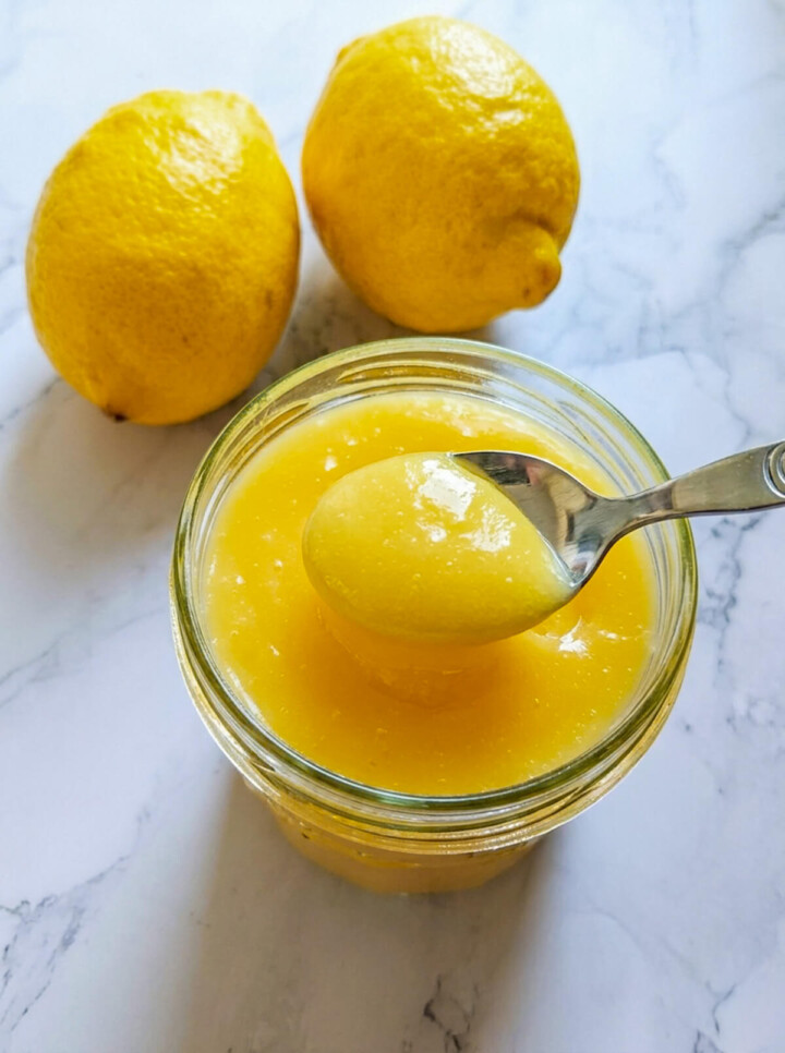 De la crème de citron (lemon curd) dans un pot de confiture en verre, avec une cuillère et deux citrons.