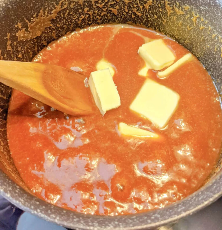 Gros plan sur la préparation d'un caramel au beurre salé maison dans une casserole, avec des morceaux de beurre frais fondant dans un sucre fondu ambré et une spatule en bois.