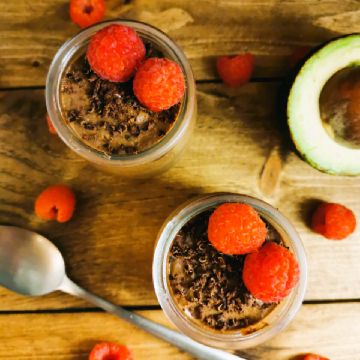 Deux petits pots en verre remplis de mousse chocolat avec framboises, cuillère sur fond en bois.