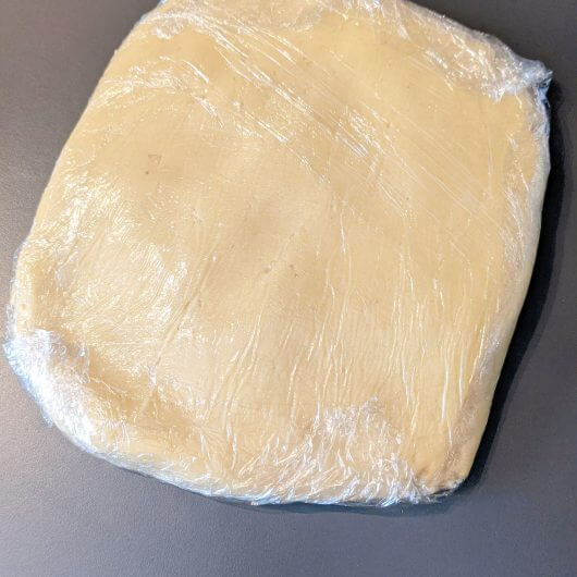 Pâton de pâte sucrée maison aplati et enveloppé dans du film alimentaire, prêt à être placé au réfrigérateur pour le temps de repos.