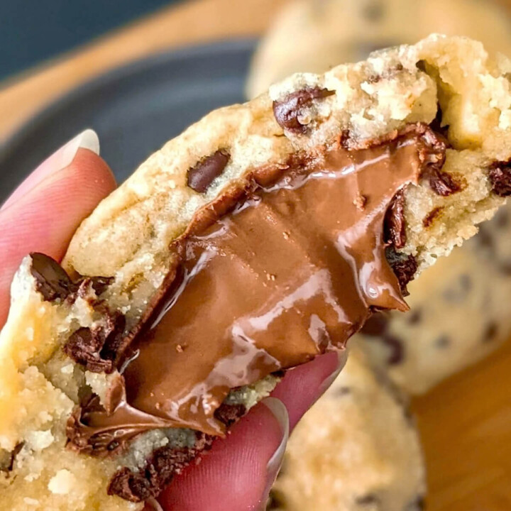 Une photo en gros plan montre un cookie aux pépites de chocolat coupé en deux, tenu par des doigts, révélant un cœur fondant de Nutella.