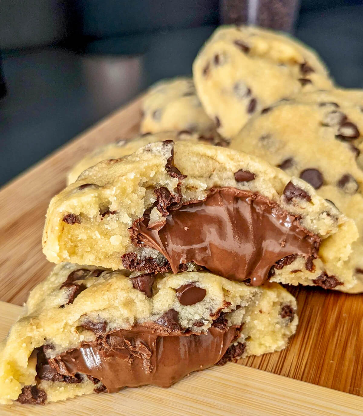 Vue rapprochée de cookies empilés sur une planche en bois, mettant en avant un cookie coupé en deux avec un cœur de Nutella très coulant et des pépites de chocolat.