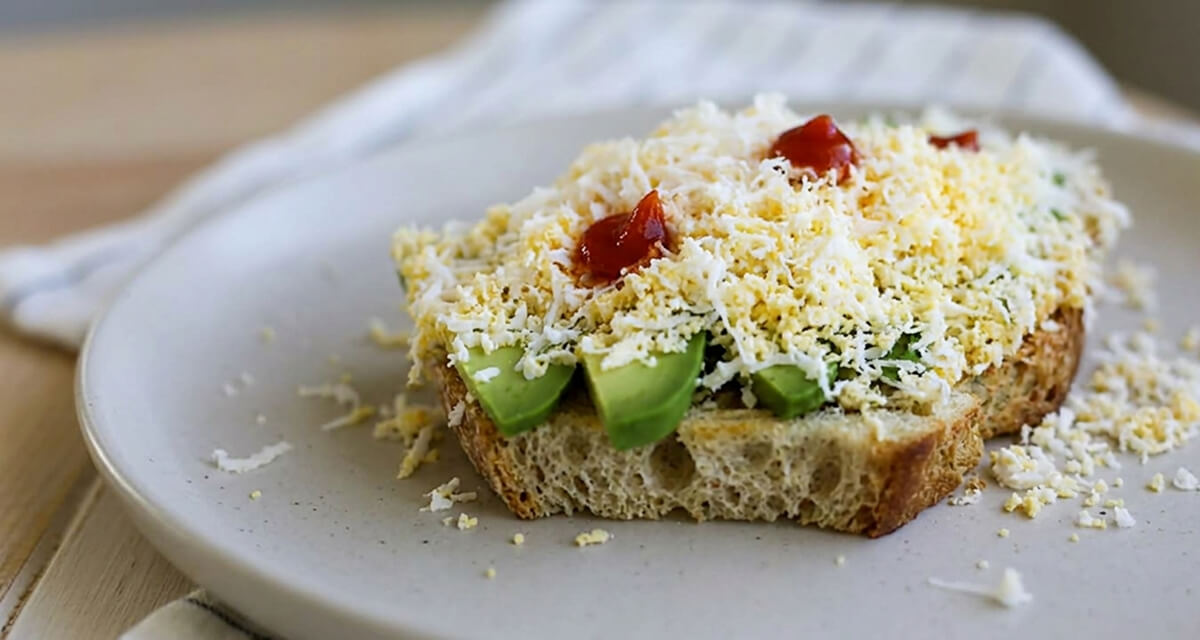 Tranche de pain de campagne grillée garnie de lamelles d'avocat frais, surmontée d'œuf dur râpé et de pointes de sauce sriracha, servie sur une assiette claire.