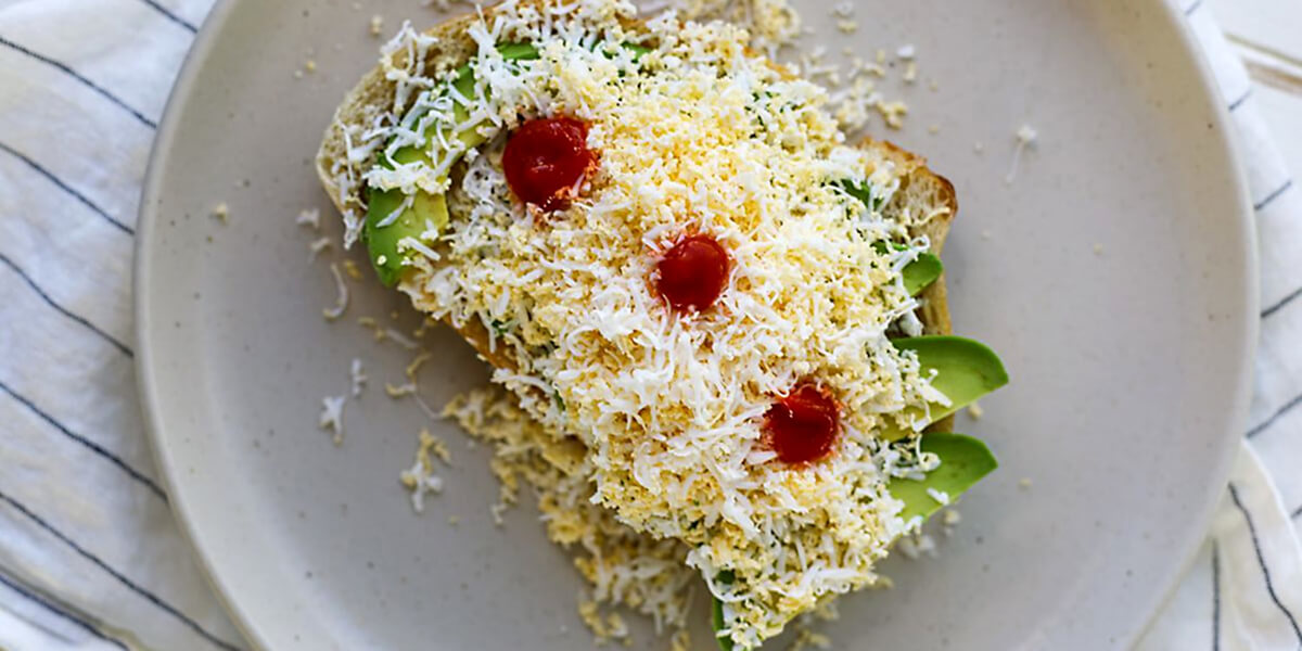 Tartine de pain grillé garnie de tranches d'avocat frais, recouverte d'un œuf dur râpé façon "mimosa" et décorée de trois pointes de sauce rouge (sriracha), présentée sur une assiette blanche. Recette virale TikTok de l'avocado toast à l'œuf râpé.