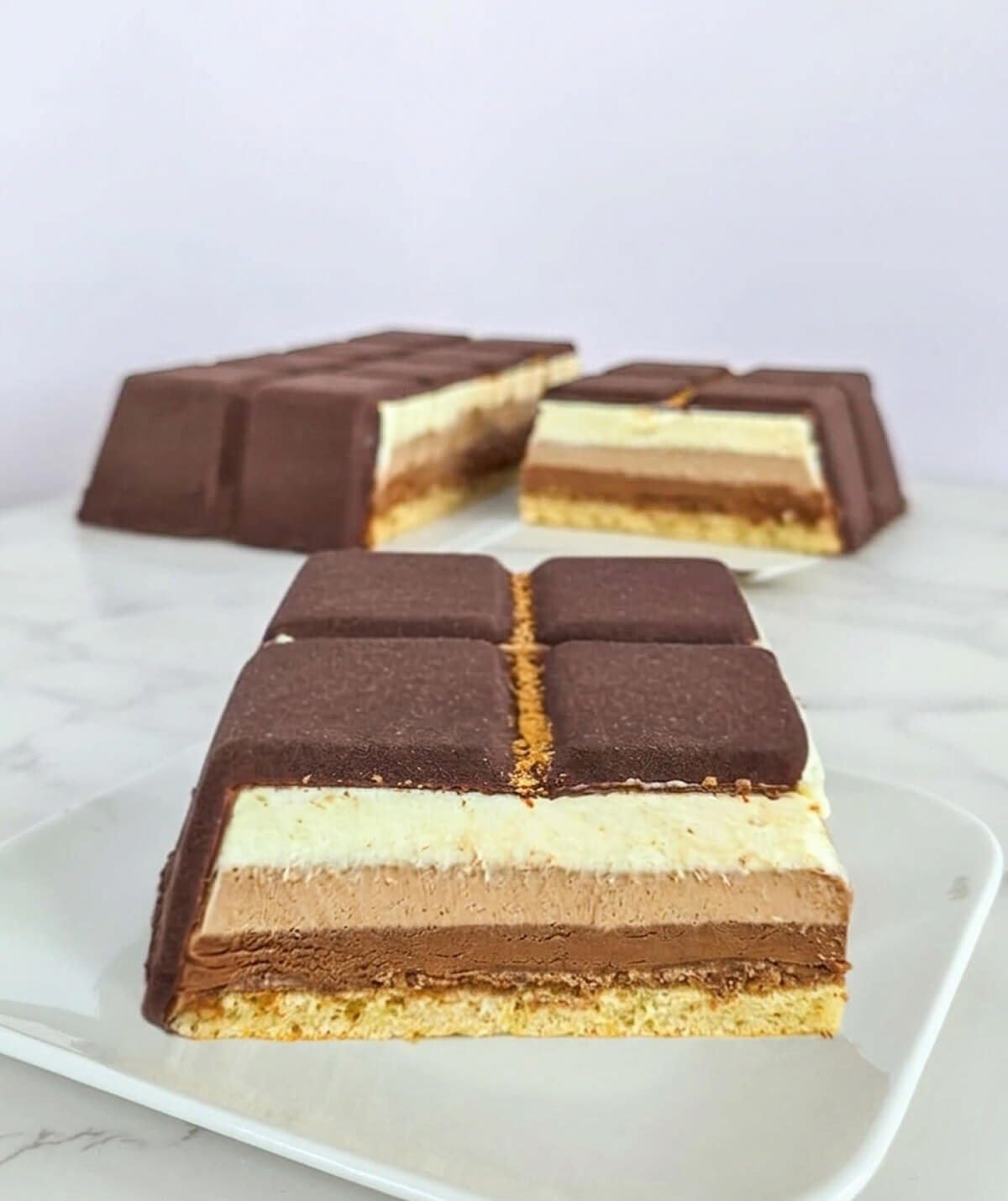Tranche d'entremets aux trois chocolats avec des couches distinctes de mousse chocolat noir, lait et blanc sur une base de biscuit Joconde moelleux.