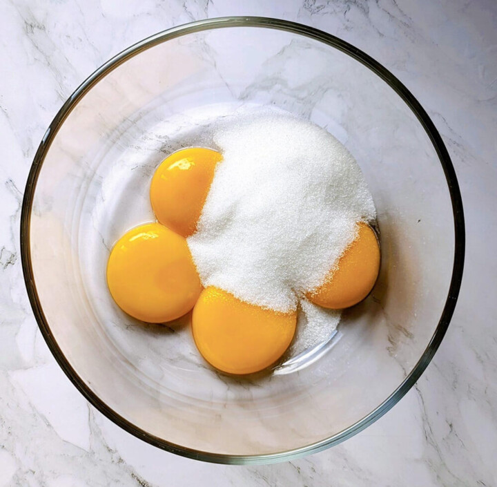 Jaunes d'œufs frais recouverts de sucre blanc dans un bol en verre pour préparer une crème anglaise maison.