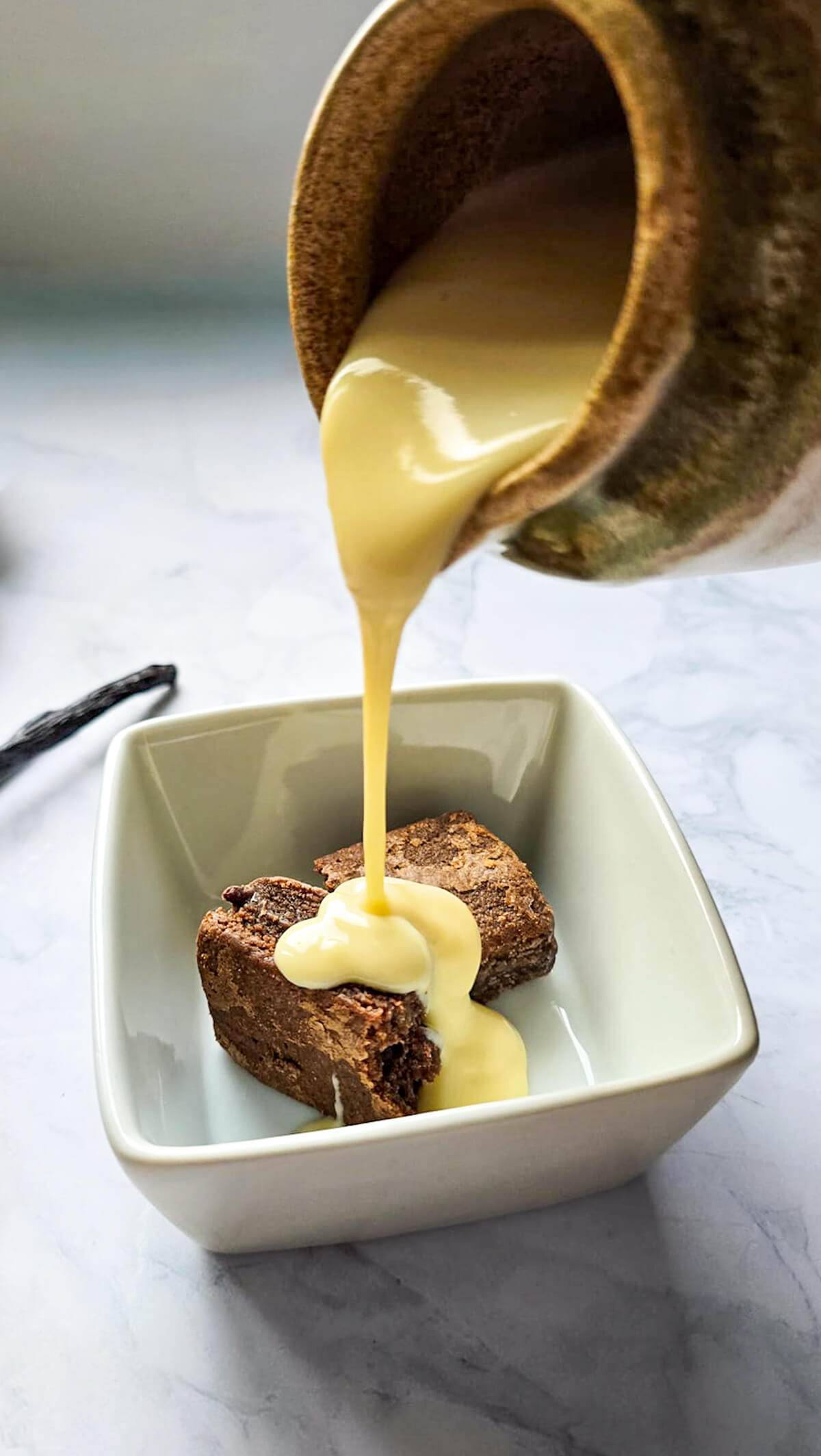 Crème anglaise onctueuse versée délicatement sur des morceaux de brownies au chocolat dans un bol blanc.