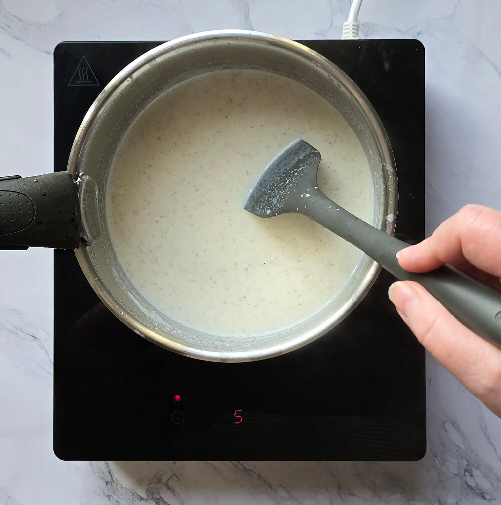 Main mélangeant progressivement du lait au roux avec une spatule grise dans une casserole, créant une sauce béchamel lisse et onctueuse.