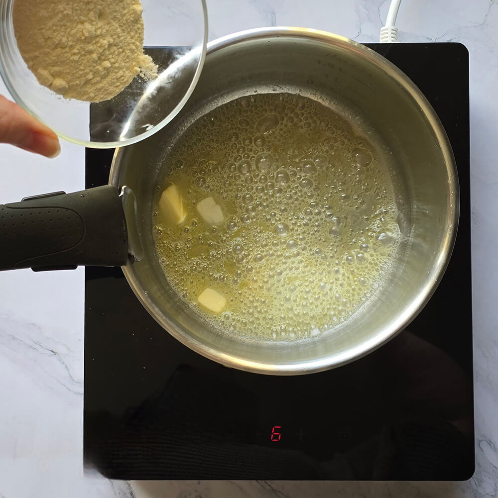 Une main versant un bol de farine dans une casserole de beurre fondu et moussant sur une plaque de cuisson à induction, étape initiale de la préparation d'un roux pour une béchamel maison.