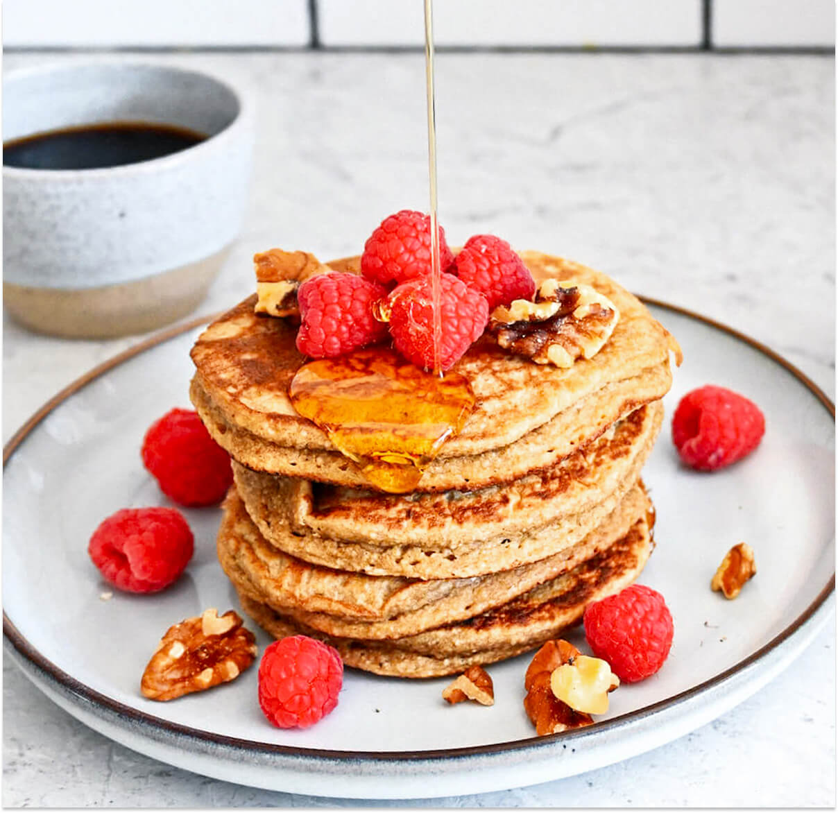 Des pancakes avoine banane dans une assiette beige, avec un filet de sirop d'érable et des framboises fraiches.
