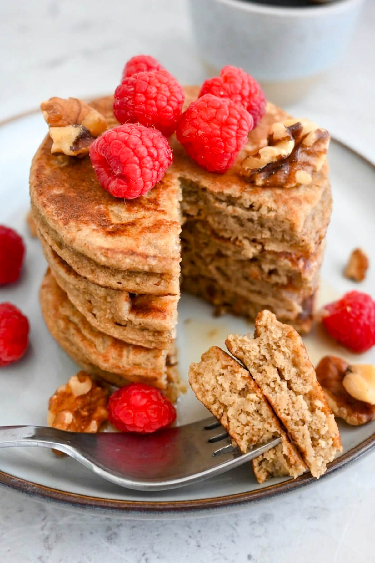 Des pancakes banane avoine healthy, dans une assiette beige, avec une fourchette tenant un morceau de pancake, framboises sur le côté.