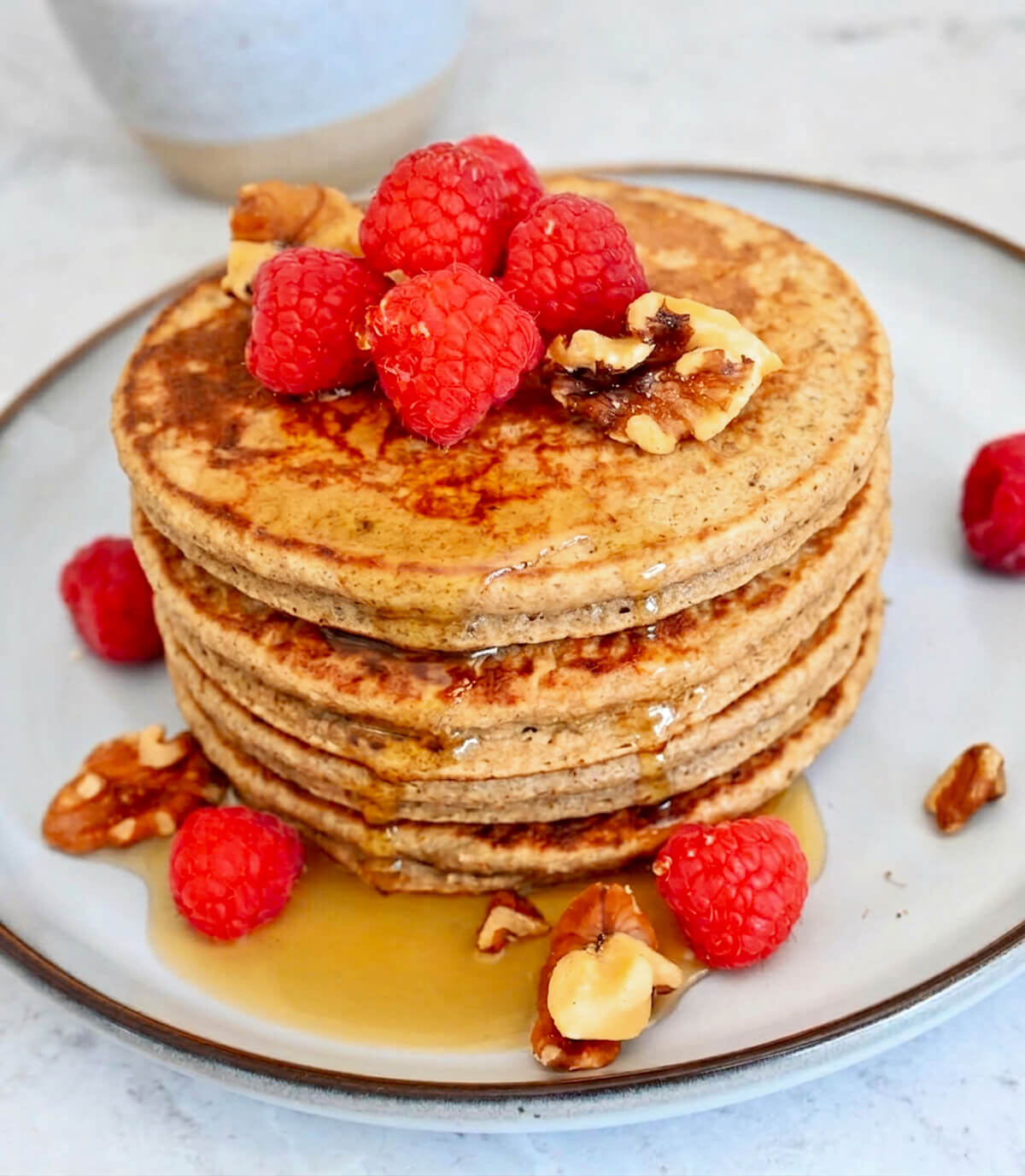 Des pancakes banane à l'avoine sur une assiette beige claire, framboises et sirop d'érable.