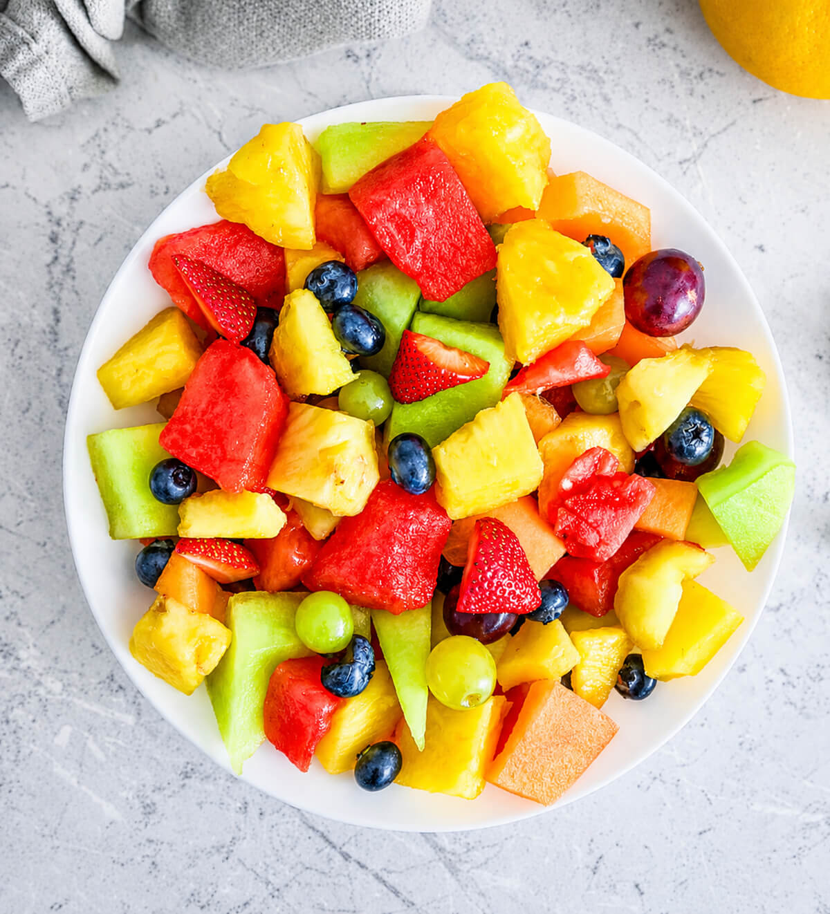 Salade de fruits frais maison vue du dessus avec pastèque, ananas, melon, fraises, myrtilles et raisins dans une assiette blanche, idéale pour un dessert léger et rafraîchissant.
