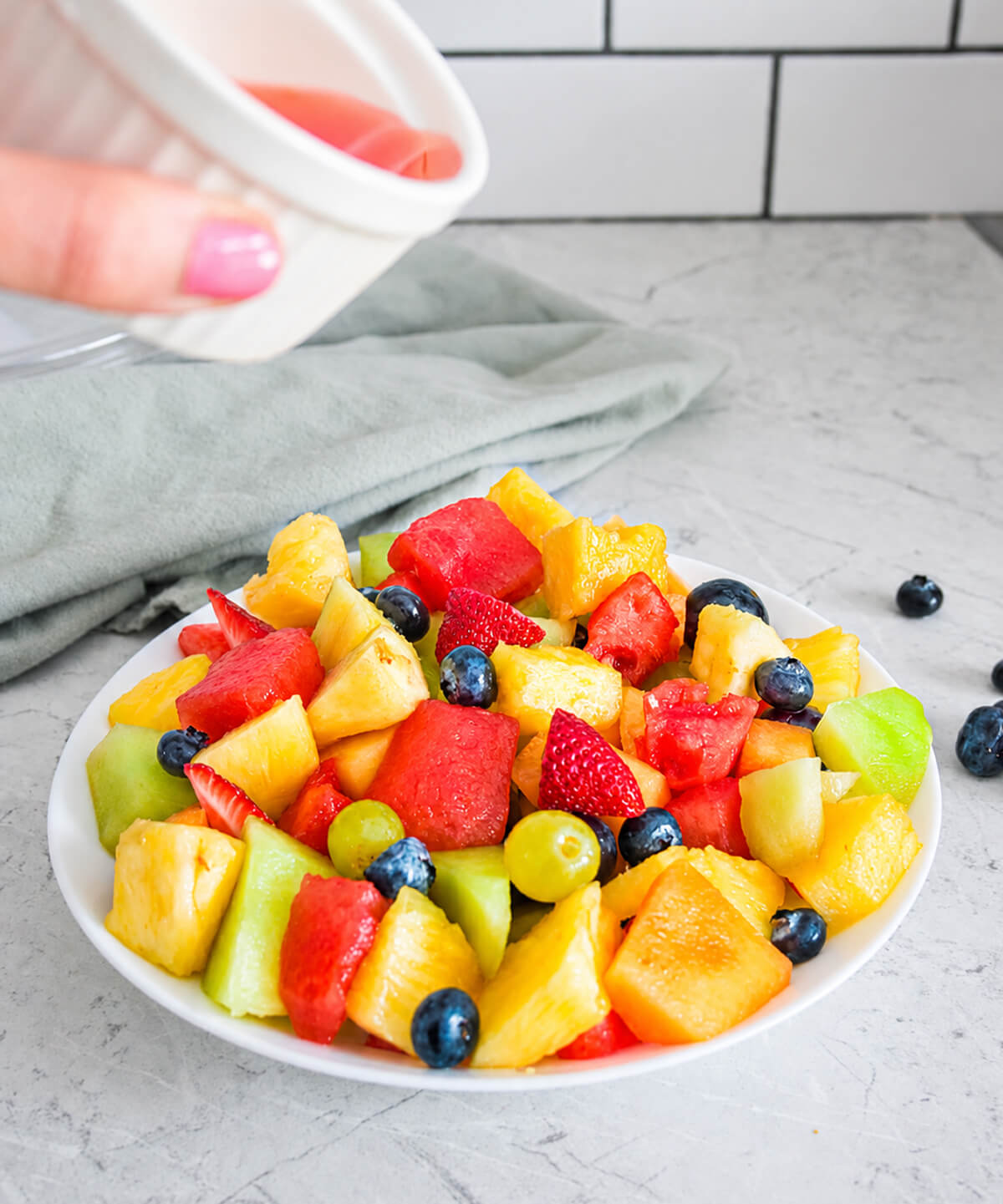Salade de fruits frais colorée servie dans une assiette blanche, composée de pastèque, ananas, melon, fraises, myrtilles et raisins, sur un plan de travail clair avec une lumière naturelle douce.