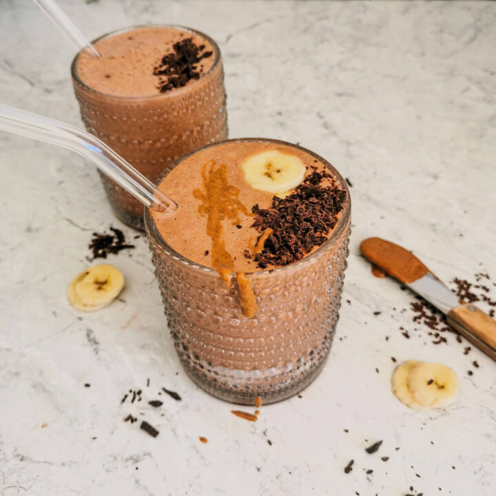 Deux verres Un verre remplis de smoothie chocolat banane, avec une paille en verre, des copeaux de chocolat, rondelles de bananes et beurre de cacahuètes.