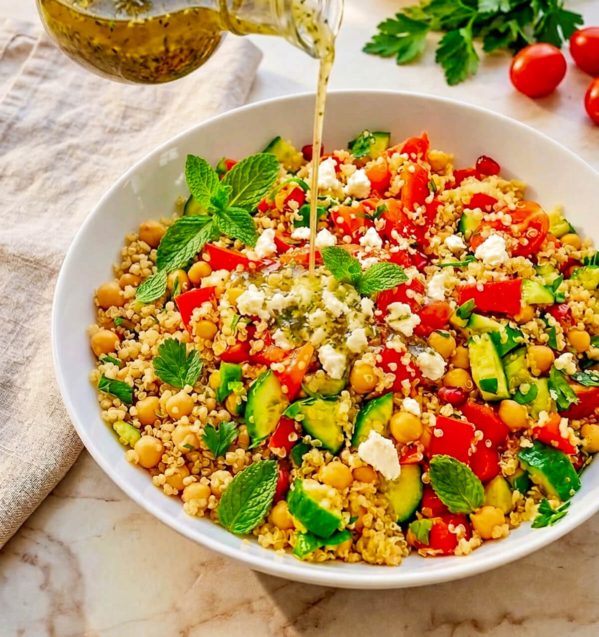 ne vinaigrette versée d'une bouteille en verre dans un bol de salade de quinoa, pois chiches, légumes et féta. La salade est présentée dans un grand bol blanc sur une surface de marbre, avec des herbes fraîches et des tomates cerises à côté.