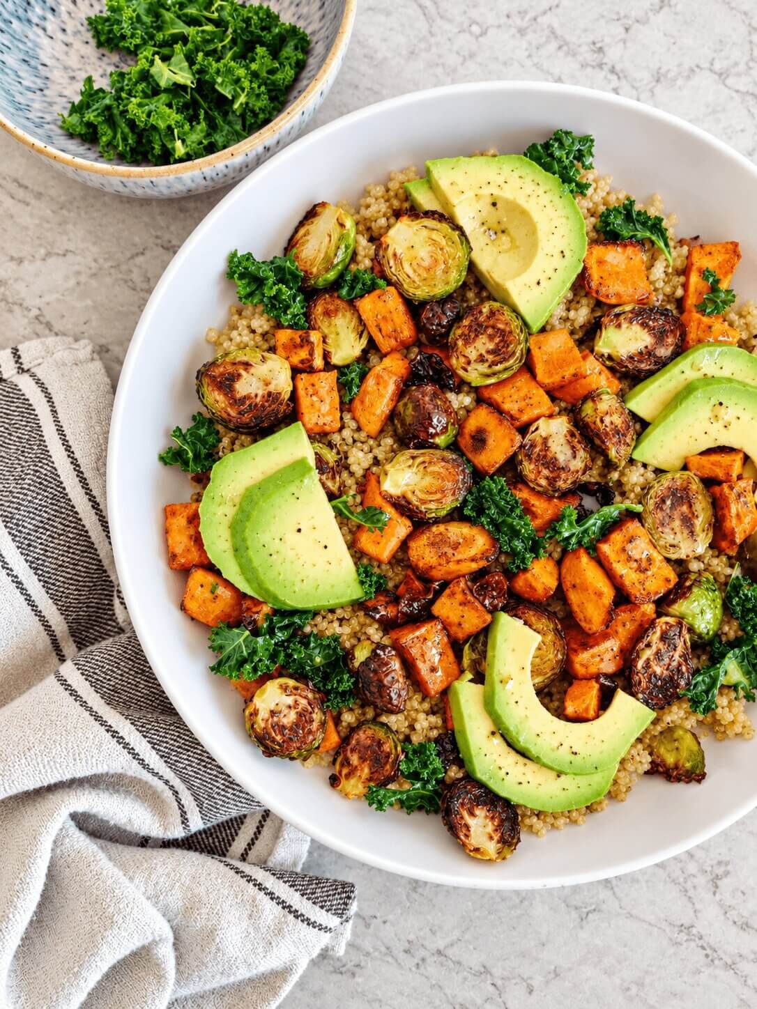 Salade de quinoa colorée dans un grand bol blanc, garnie de patates douces rôties, choux de Bruxelles grillés, chou kale et tranches d’avocat, servie sur un plan de travail en marbre clair.