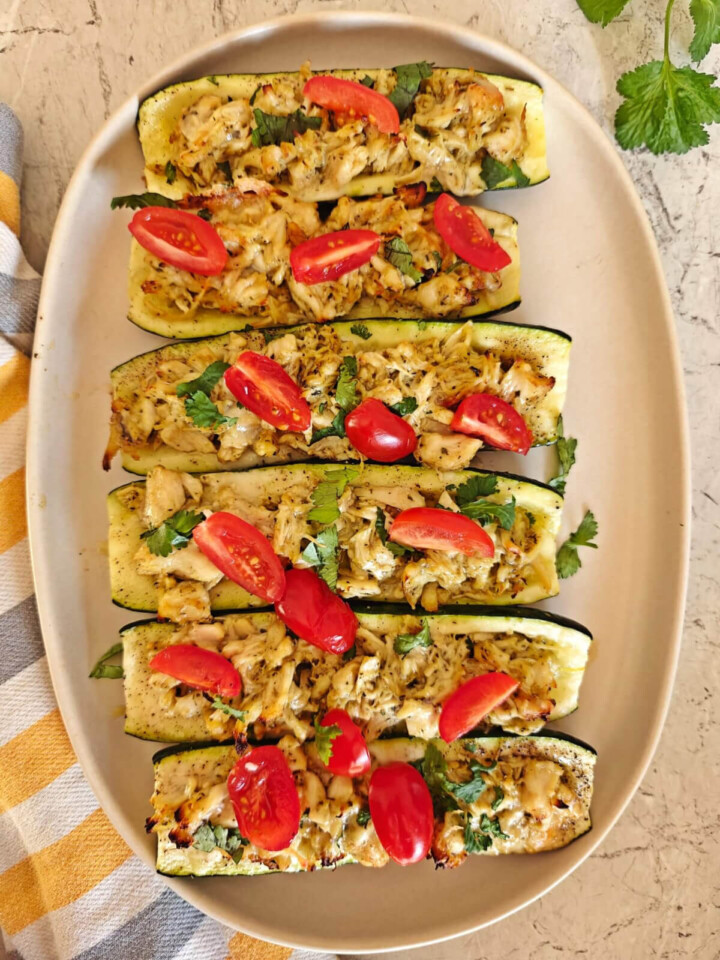 Des courgettes farcies au poulet pesto dans un play beige avec un torchon gris et jaune sur le coté