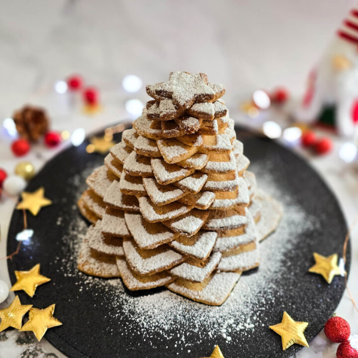 Un sapin de Noel en biscuits étoilés recouvert de sucre glace blanc, sur une assiette noire avec des décorations de Noel sur un fond blanc marbré.