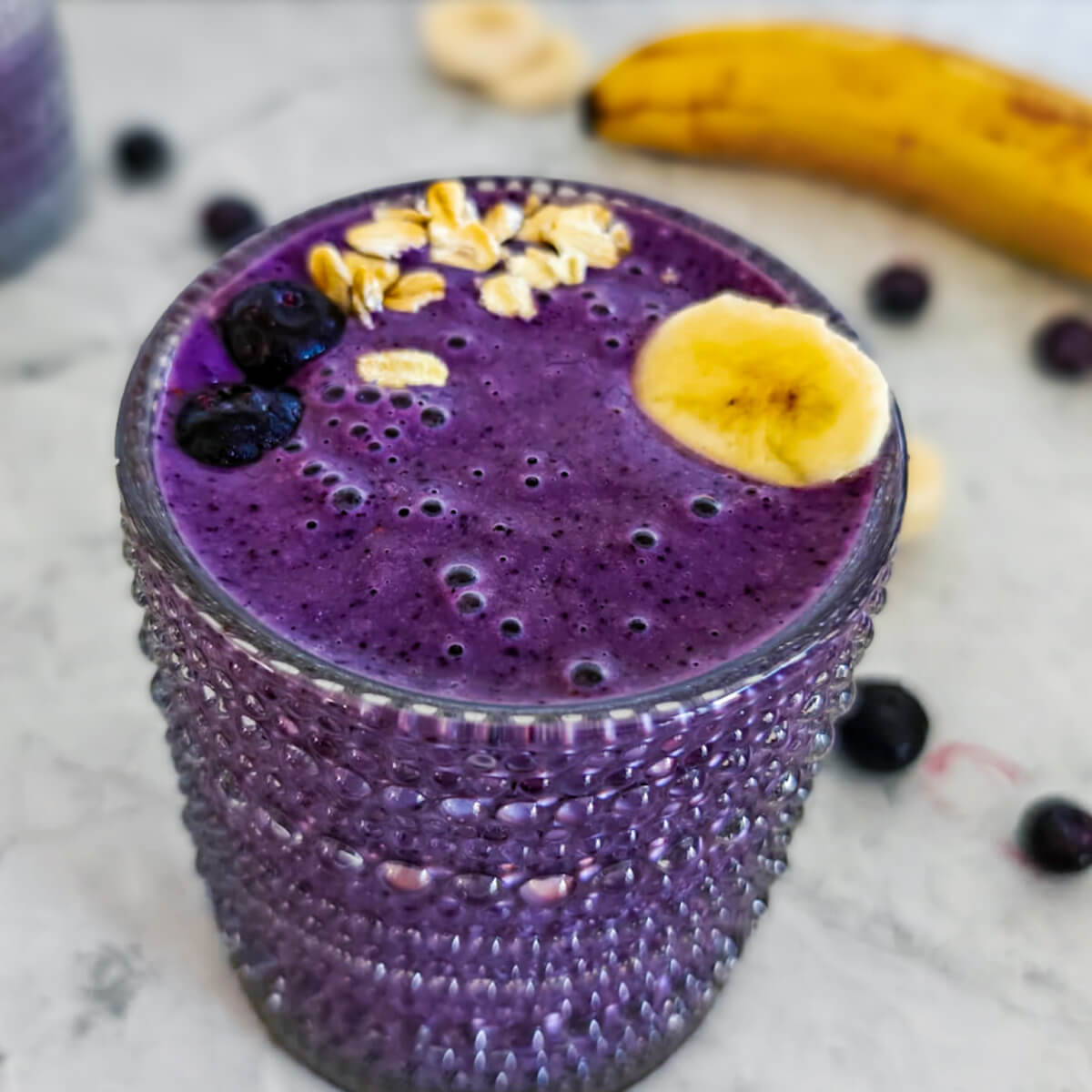 Un verre transparent rempli de smoothie aux myrtilles avec des myrtilles et rondelles de bananes sur un fond gris clair.
