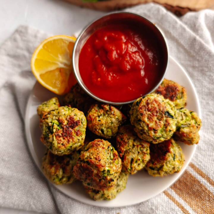 Des bouchées de courgettes apéro dans une assiette blanche avec un morceau de citron et de la sauce tomate dans un petit pot.