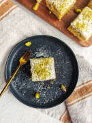 Un morceau de gâteau pistache dans une assiette noire, torchon beige dessous l'assiette.