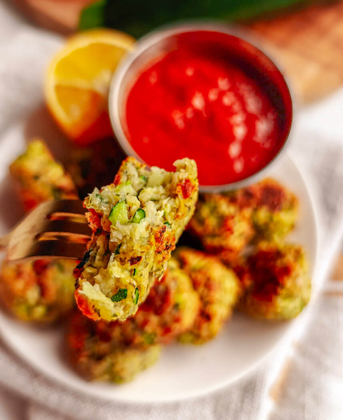 Une fourchette tenant un morceau de bouchée de courgettes avec au fond, une assiette remplie de bouchées de courgettes.