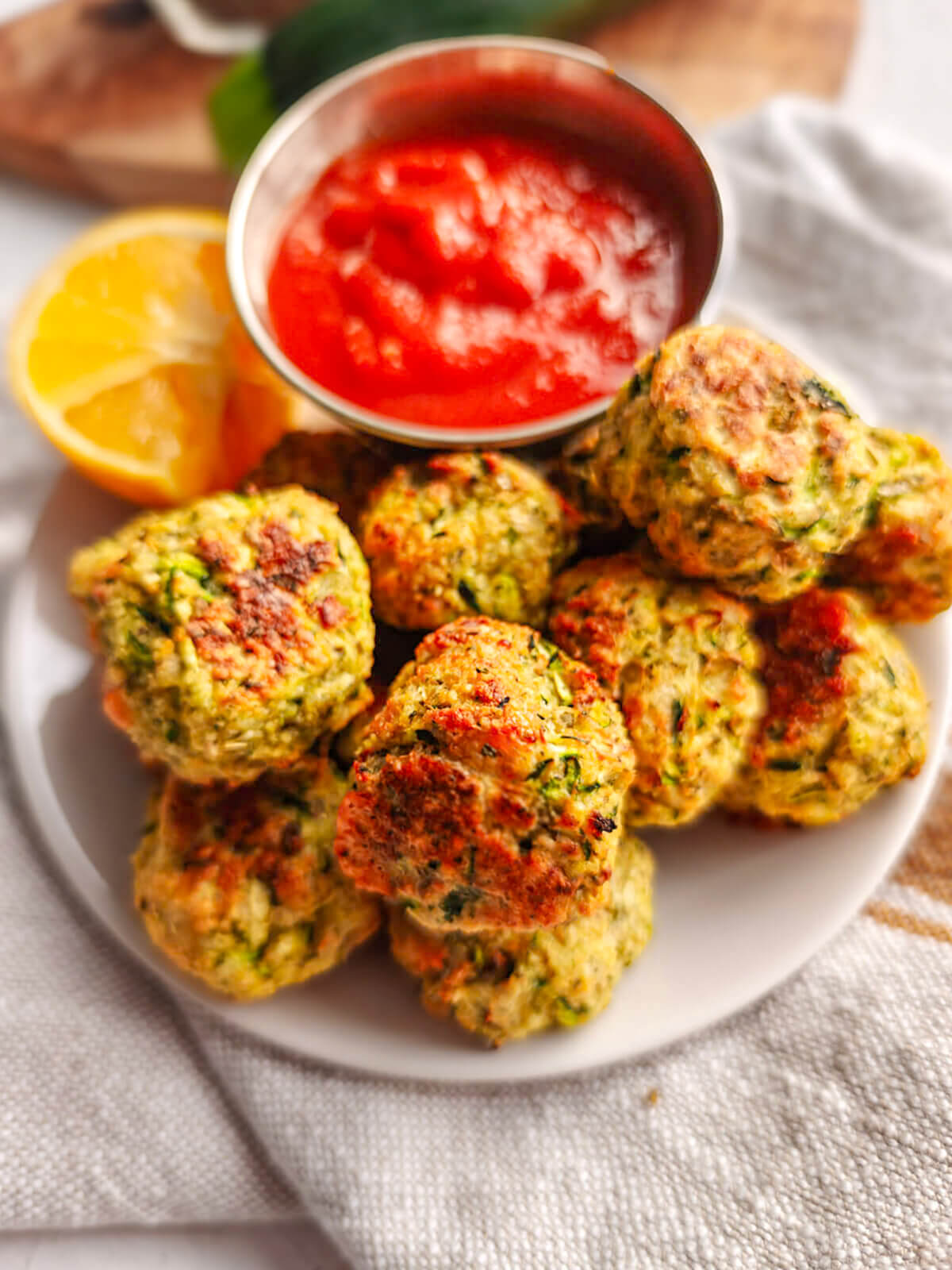 Des bouchées de courgettes parmesan sur une assiette blanche avec un bout de citron et une sauce tomate.