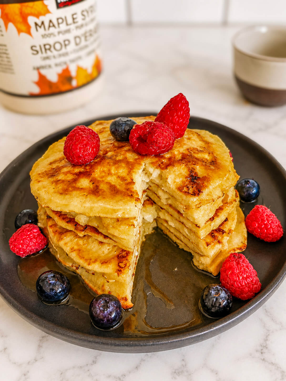 Une pile de pancakes protéinés, moelleux et dorés sur une assiette sombre, garnis de framboises fraîches et de myrtilles, nappés de sirop d'érable avec une bouteille de sirop d'érable en arrière-plan.