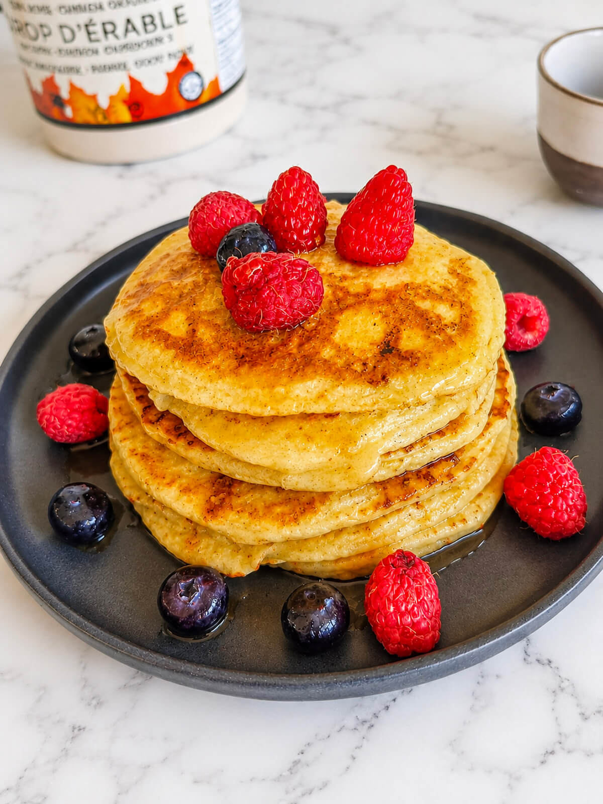 Des pancakes protéinés sans whey, bien fluffy, empilés les uns sur les autres, framboises sur le dessus et côté, sur une assiette noire, sur fond blanc marbré