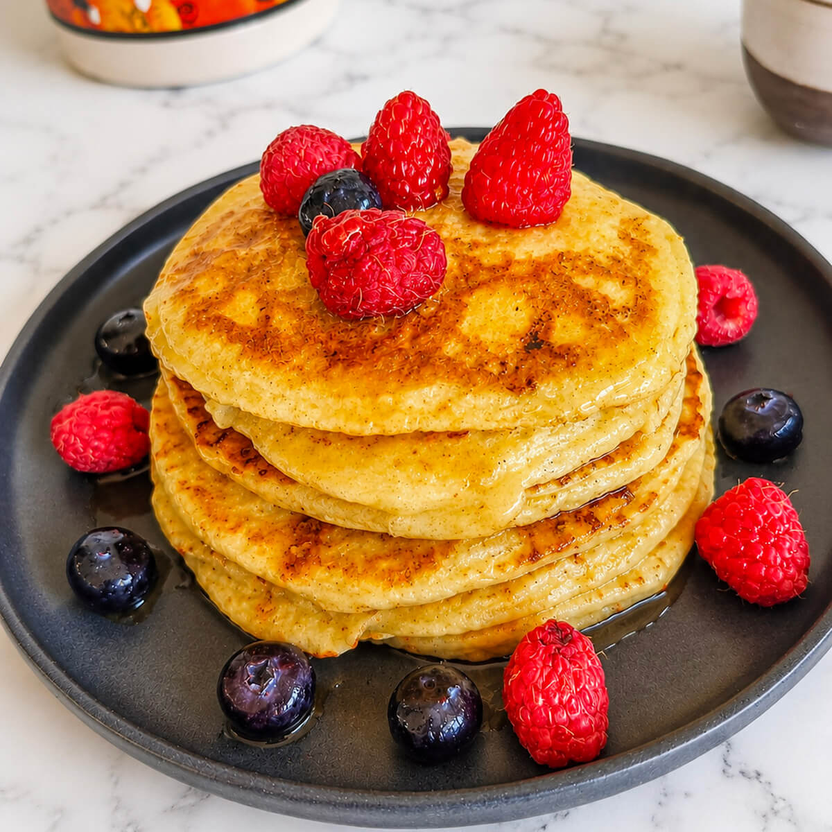 Des pancakes protéinés sans whey, bien fluffy, empilés les uns sur les autres, framboises sur le dessus et côté, sur une assiette noire, sur fond blanc marbré.
