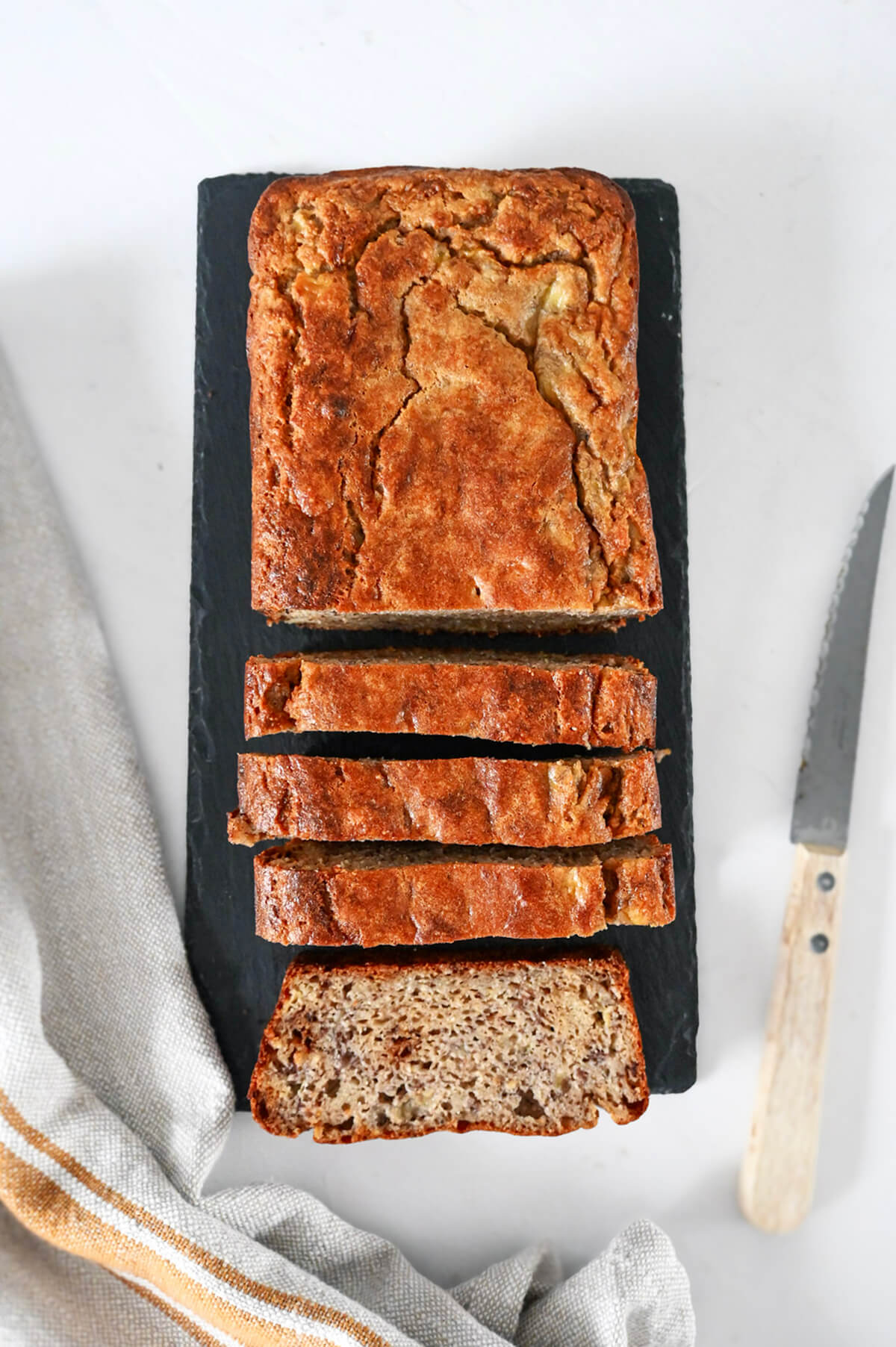 Des tranches de banana bread sur une planche noire, couteau et torchon sur le coté.