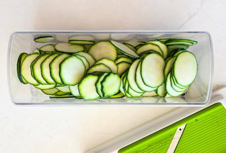 Des rondelles de courgettes dans le bac d'une mandonline, sur un fond blanc.