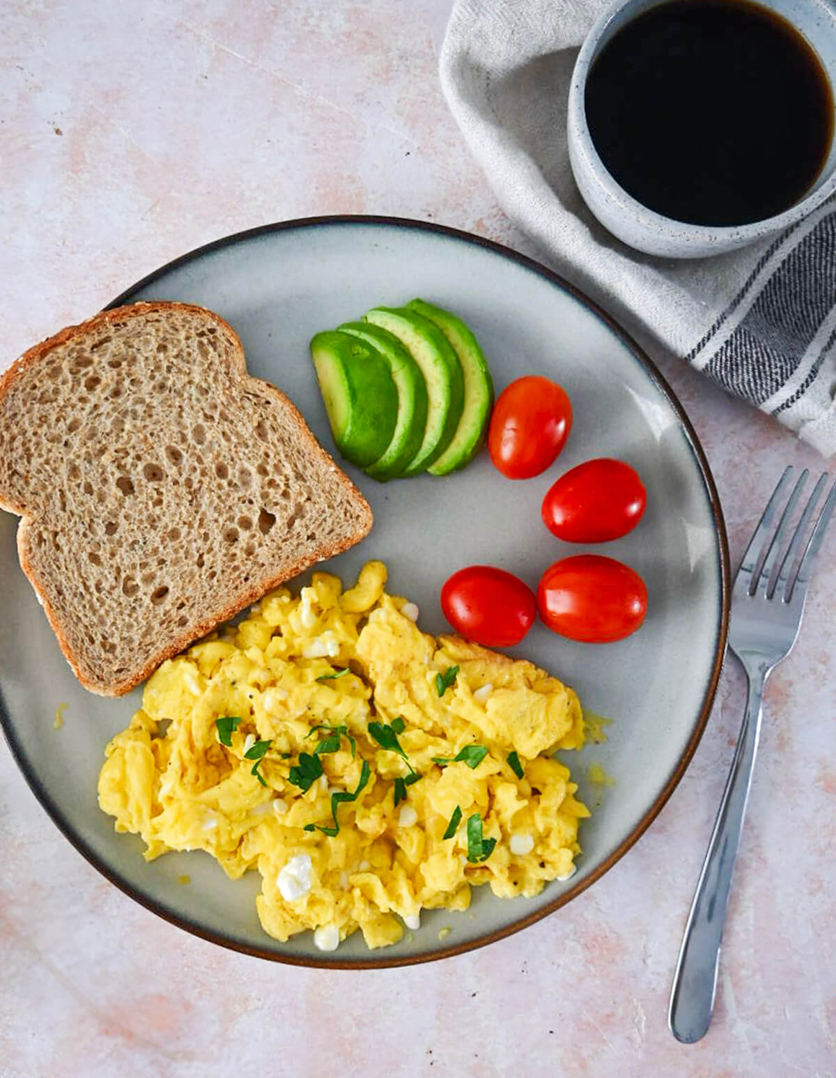 Des oeufs brouillés avec cottage cheese avec tomate, avocat et pain sur une assiette beige, café sur le coté.