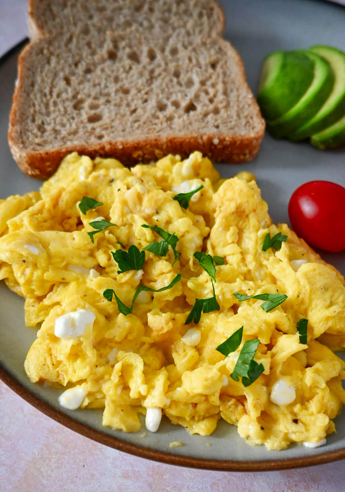 Des oeufs brouillés avec cottage cheese avec tomate, avocat et pain sur une assiette beige, café sur le coté.