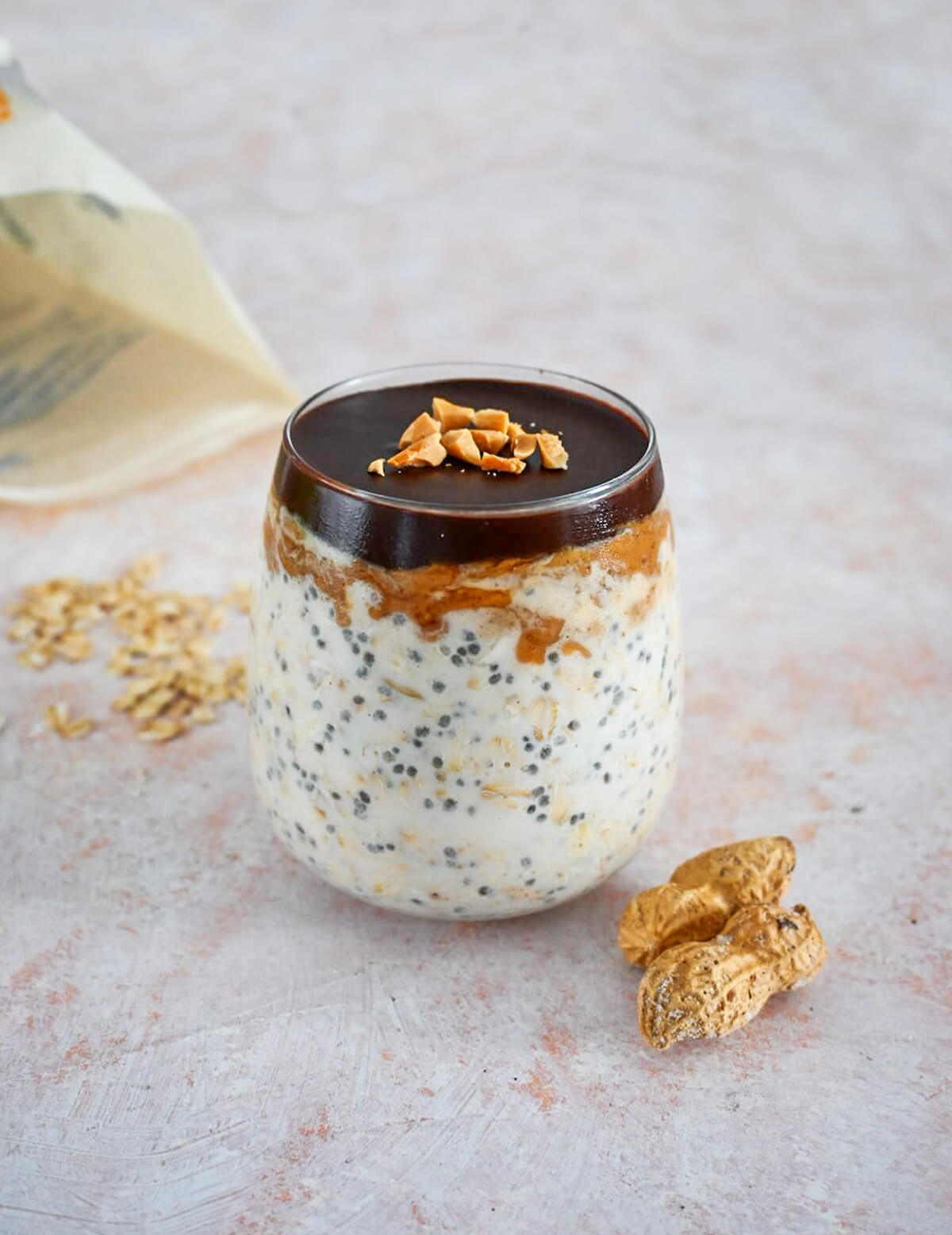 Un overnight oats snickers dans un verre sur une surface rose clair, cacahuètes et flocons d'avoines ur le coté.