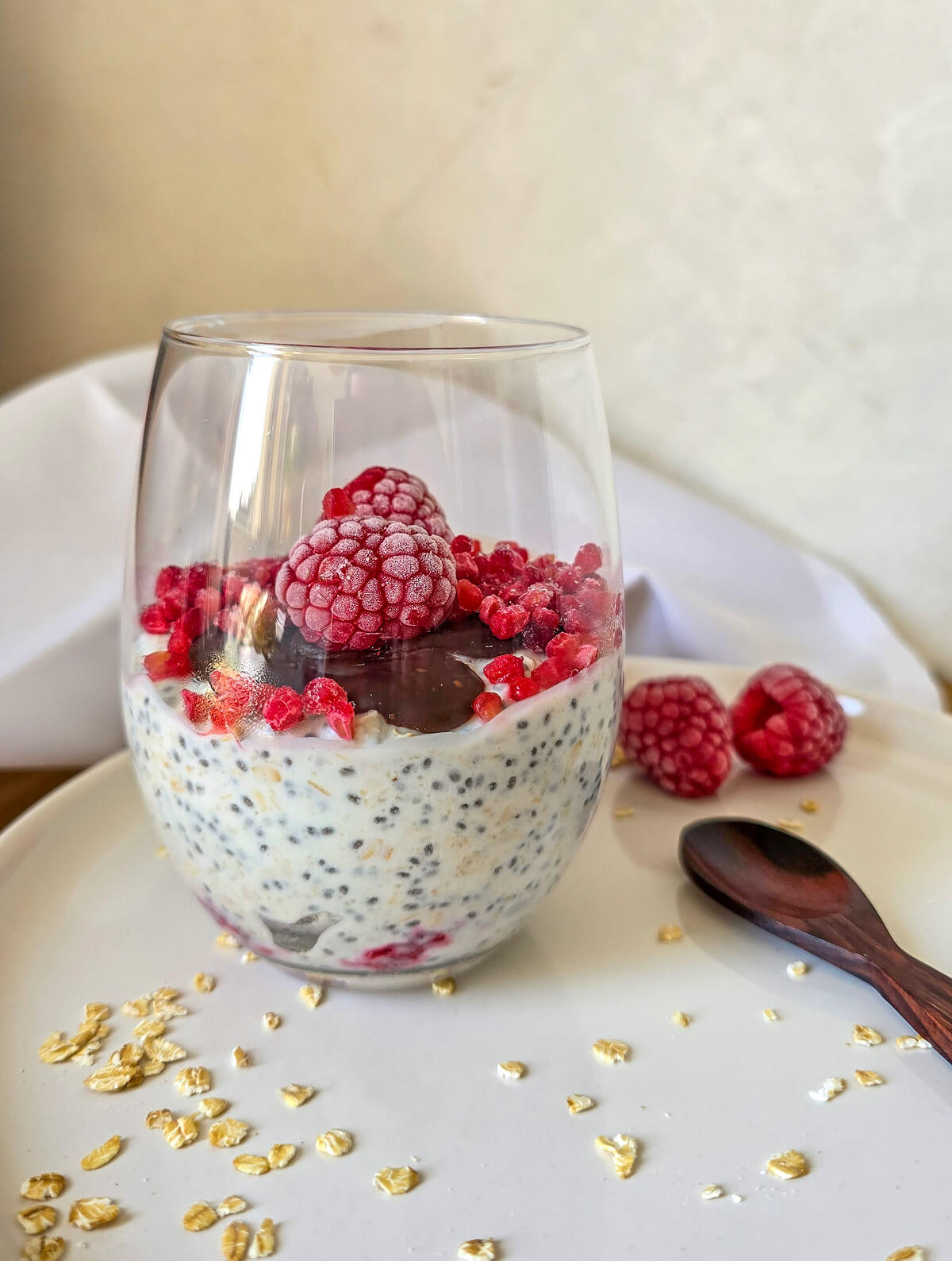 Un verre rempli d'un overnight porridge framboise, sur une assiette blanche, cuillère en bois sur le coté.