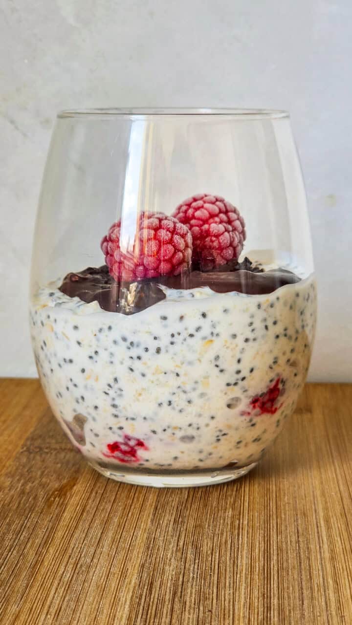 Un verre rempli d'un overnight porridge framboise, sur une assiette blanche, cuillère en bois sur le coté.