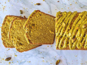 Une vue de dessus d'un cake à la pistache moelleux, coupé en tranches, révélant une mie verte tendre. Le gâteau est surmonté d'un glaçage onctueux à la pistache et parsemé de pistaches concassées sur un fond en marbre blanc.