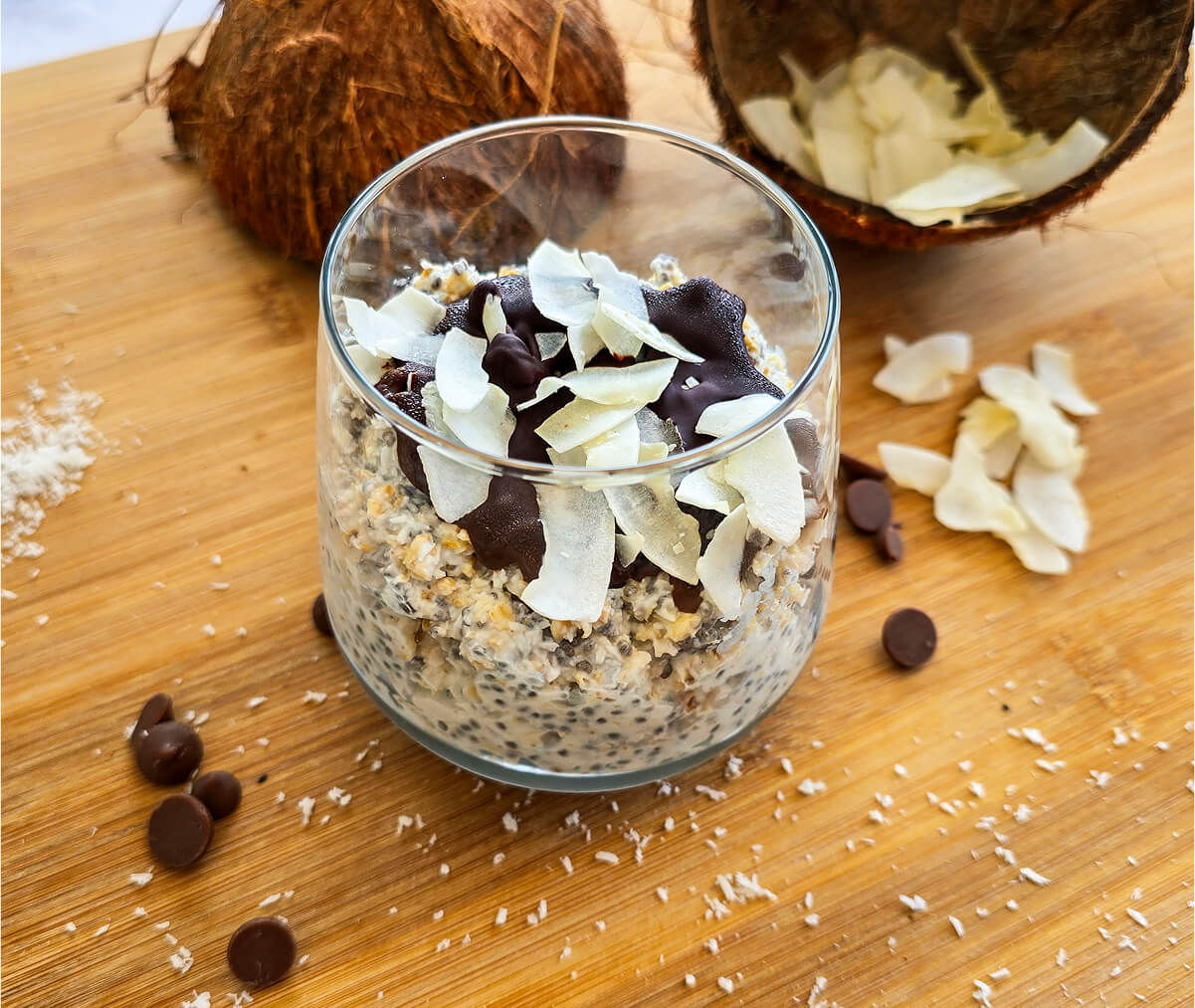 Un overnight porridge bounty dans un verre, sur une planche de bois, pépites de chocolat et copeaux de noix de coco sur le coté.