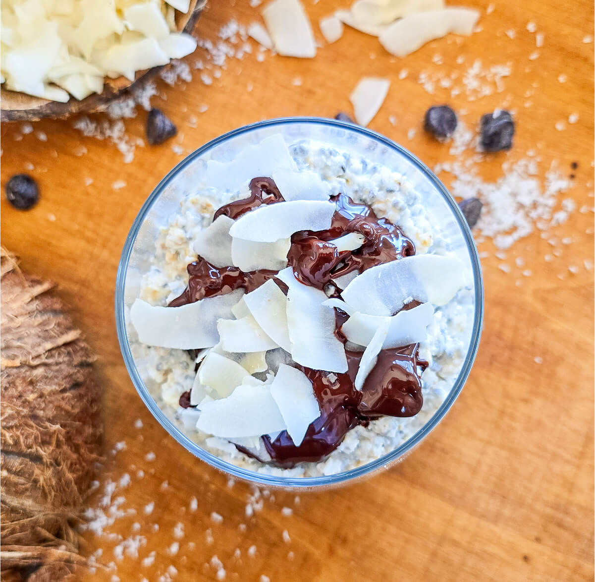 Un overnight porridge bounty dans un verre, sur une planche de bois, pépites de chocolat et copeaux de noix de coco sur le coté.