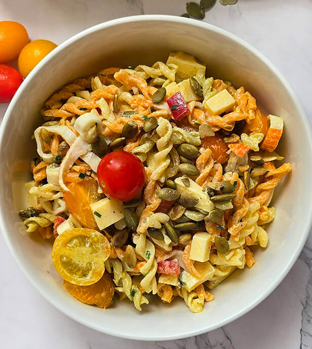 Une salade de Pâtes au Surimi sur un fond blanc marbré, tomates cerises et graines sur le coté.