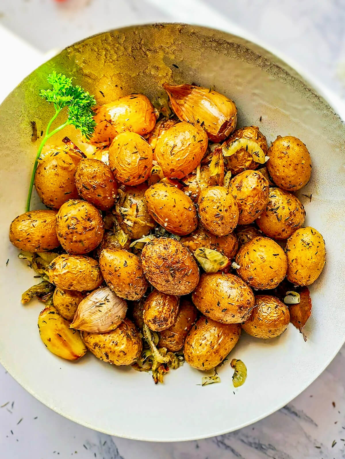 Une assiette creuse de pommes de terre grenailles rôties à l'airfryer, bien dorées avec des herbes de Provence, du persil frais et des gousses d'ail en chemise.