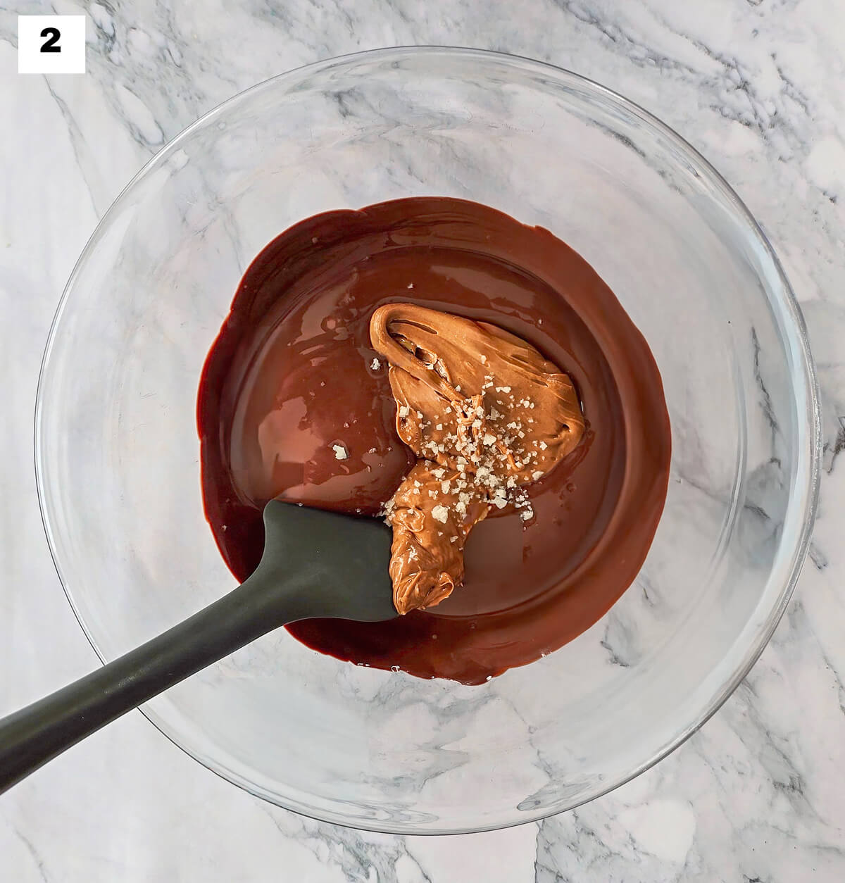 Du chocolat fondu et du praliné noisettes dans un saladier eb verre, sur un fond mabré avec une spatule.
