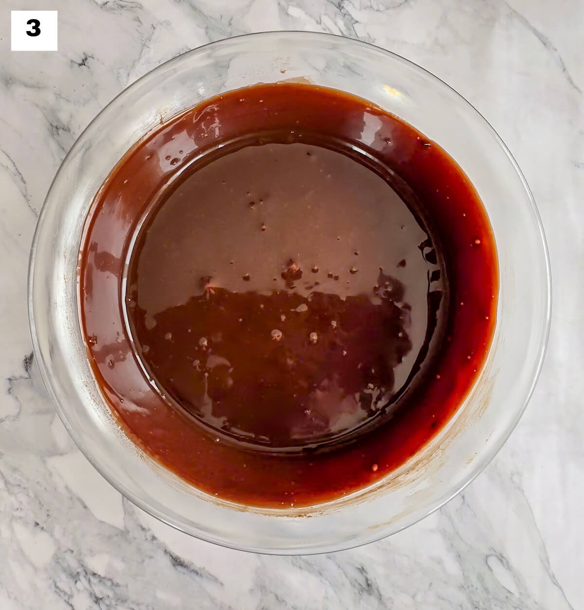De la ganache chocolat fondue dans un saladier en verre, sur un fond blanc marbré.