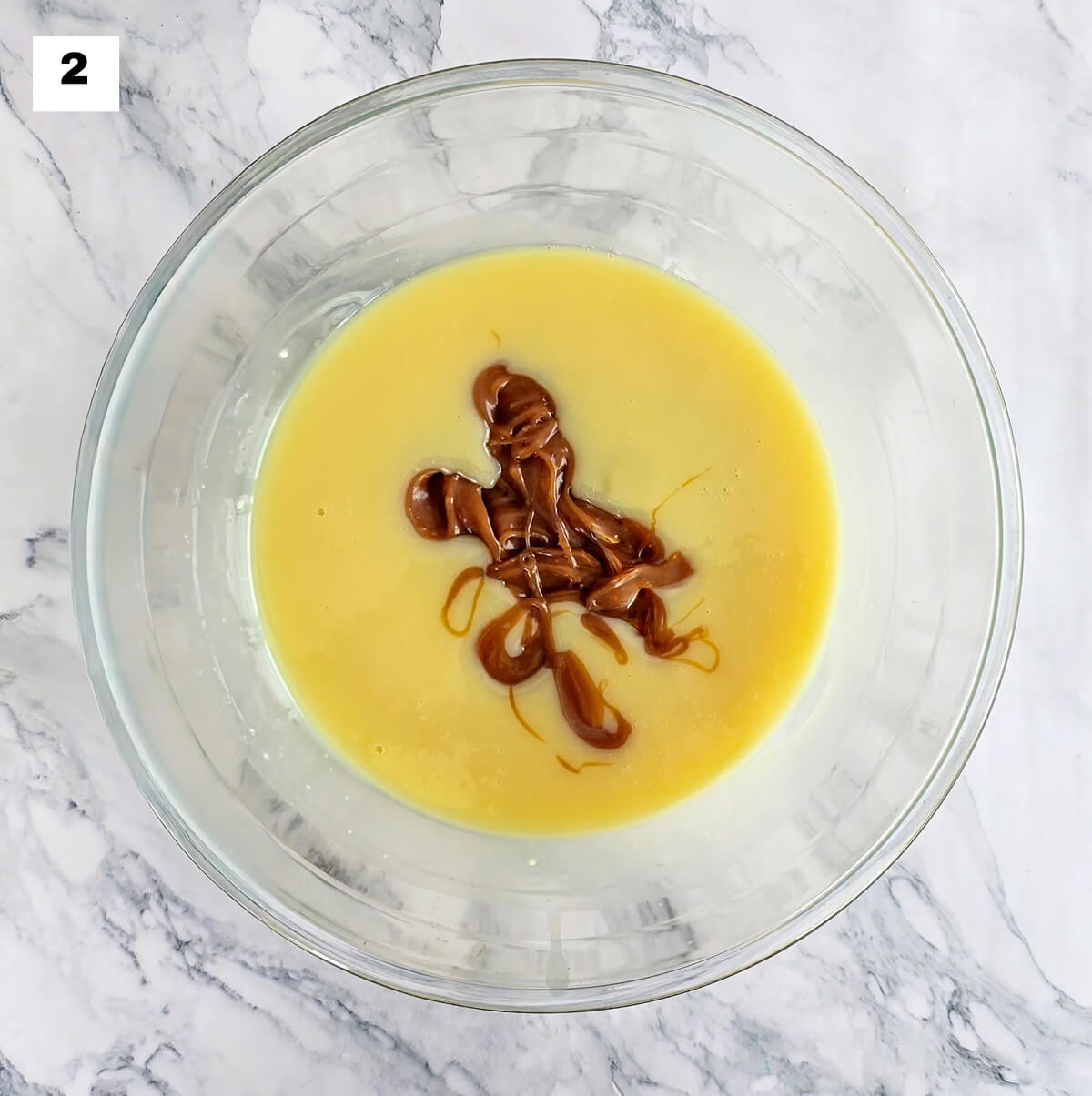 Un saladier en verre contenant du chocolat blanc fondu et du caramel beurre salé, sur un fond blanc marbré.