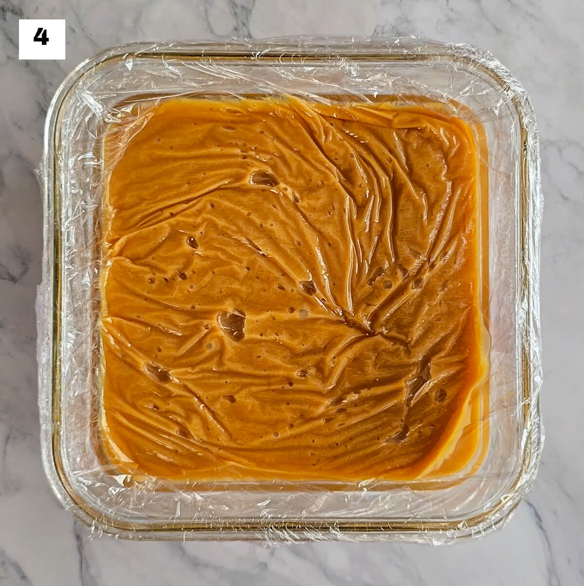 De la ganache caramel beurre salé dans une boite de conservation en verre carré, sur un fond blanc marbré.