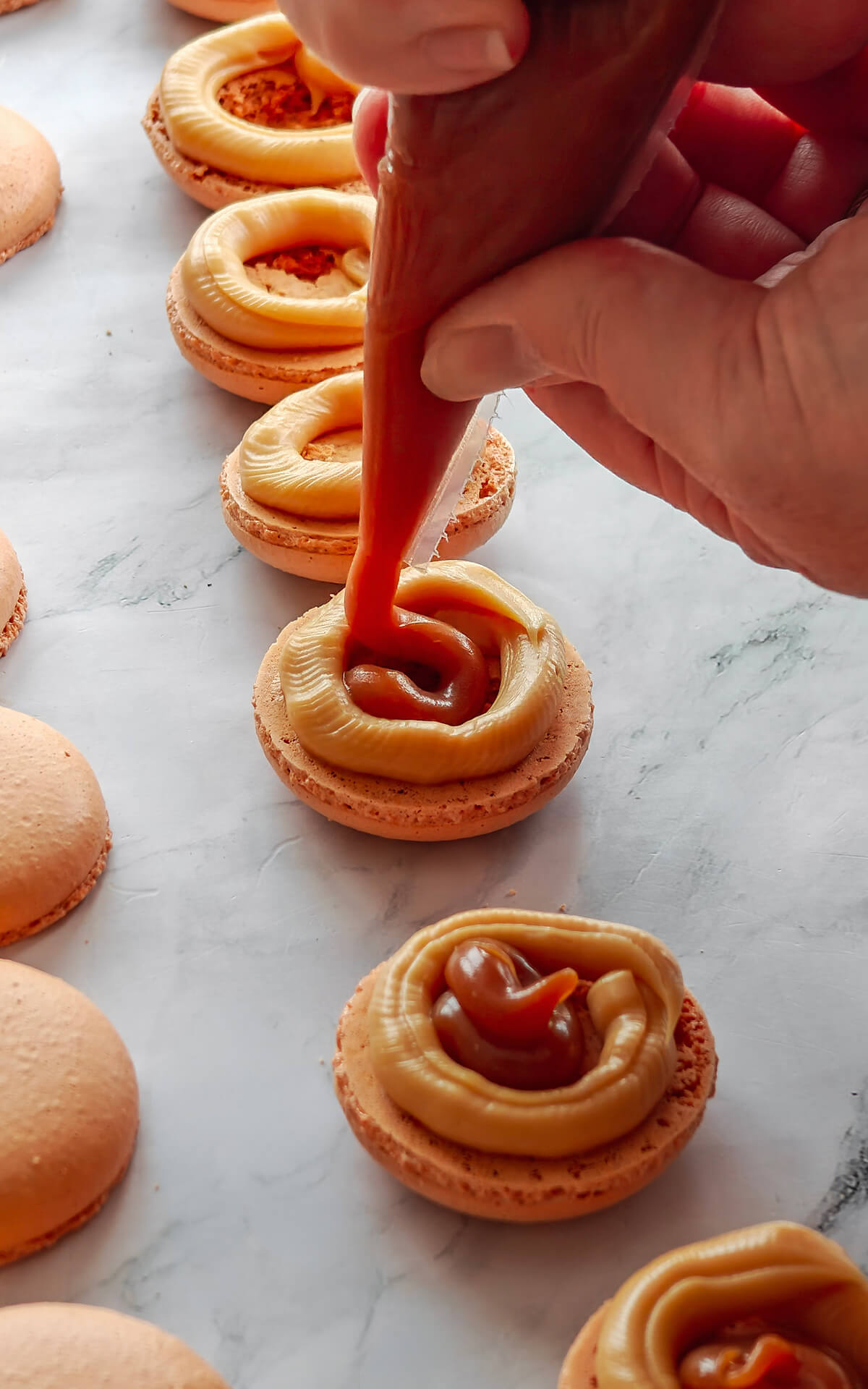 Deux mains, tenant une poche à douille, remplissent des coques de macarons avec du caramel au beurre salé.
