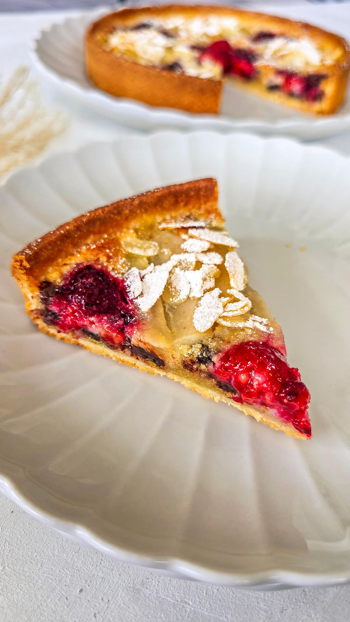 Une part de tarte amandine poire framboise dans une assiette blanche.