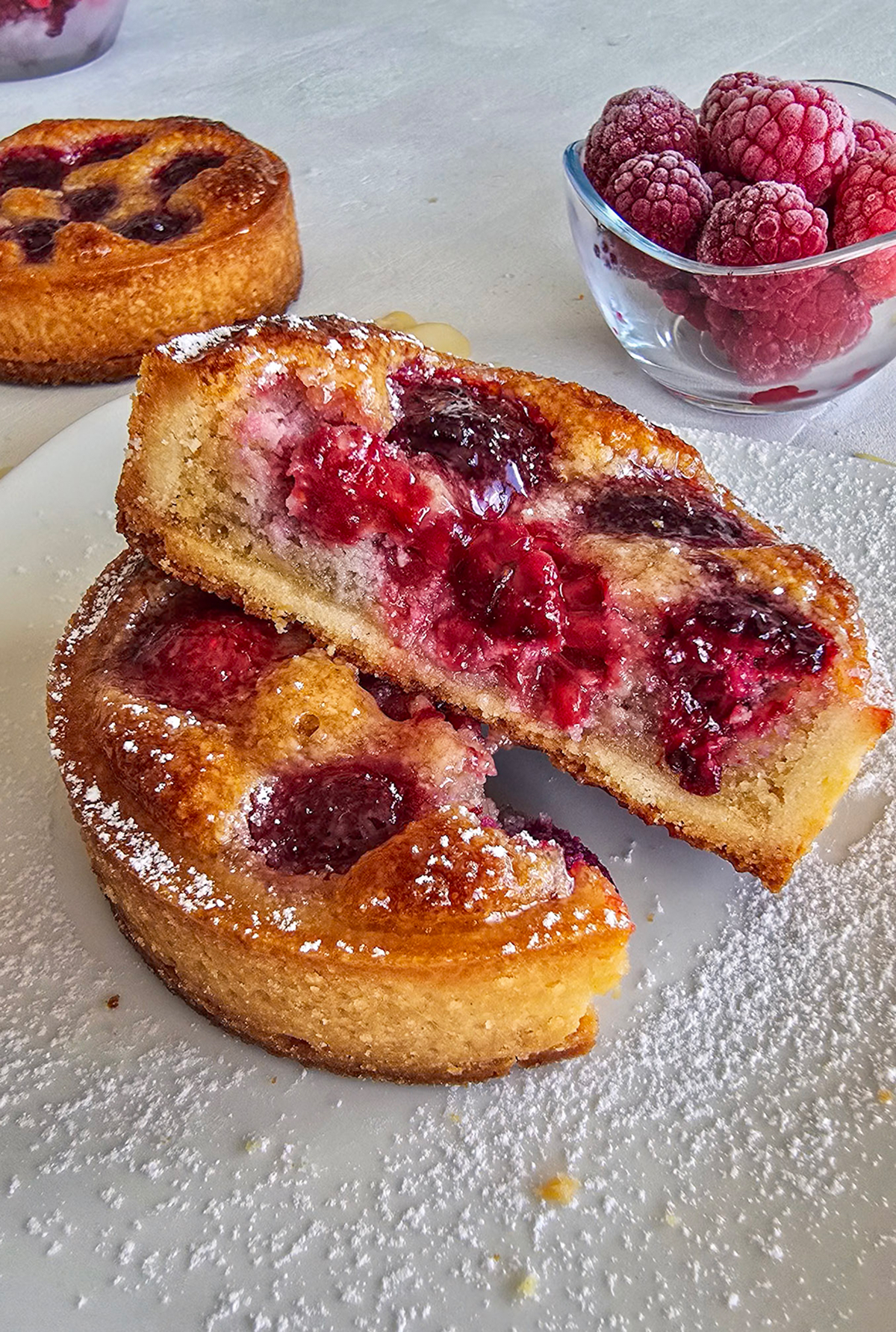 Une tartelette framboise coupé en deux, sur une assiette blanche, framboise en deuxième plan.